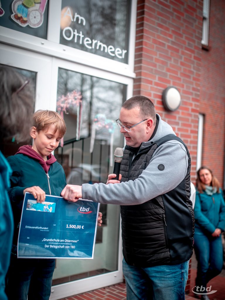 Thomas Schmidt übergibt den symbolischen Scheck im Namen der Belegschaft an Schulsprecher Leano.